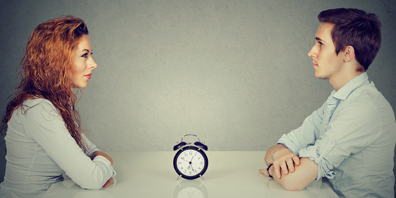 Frau und Mann sitzen einander gegenüber, in der Mitte eine Stoppuhr. Symbolbild für Speed Dating