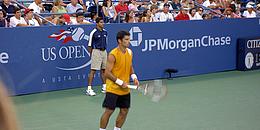 Ein Tennisspieler mit gelbem Shirt, schwarzen Shorts und weißen Schuhe häkt seinen Schläger