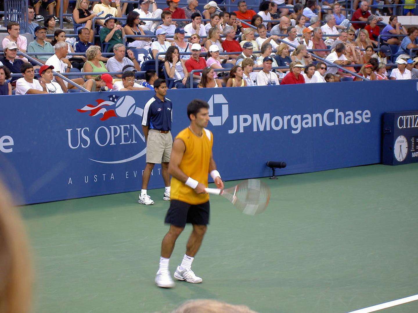 Ein Tennisspieler mit gelbem Shirt, schwarzen Shorts und weißen Schuhe häkt seinen Schläger