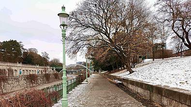 Neben einem offenen Kanal ist ein asphaltierter Weg mit Laternen und einer schneebedeckten Wiese mit Bäumen, die über den Weg ragen.