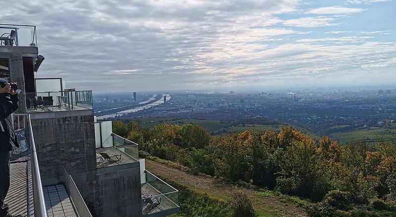 Ein Panoramablick über Wien und die Donau. Am linken Bildrand sind ein paar Menschen zu sehen, die von einem asphaltierten Bereich aus Fotos machen.