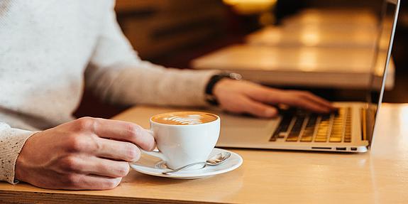 Person sitzt mit Cappucchino im Kaffeehaus und arbeitet am Laptop.