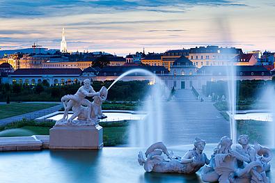 Großer Springbrunnen mit Skulpturen im Vordergrund, im Hintergrund das beleuchtete Untere Belverde abends. 