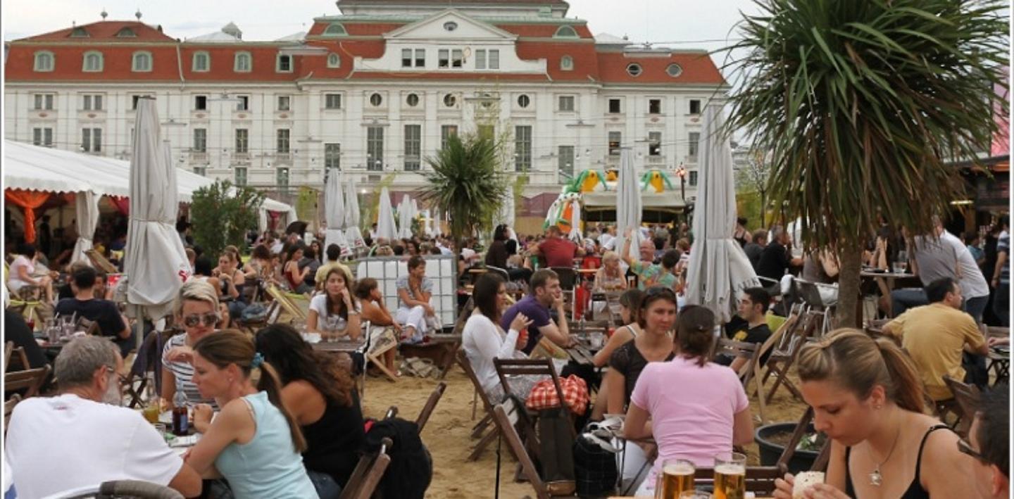 Viele Menschen sitzen draußen auf Stühlen im Sand. Im Hintergrund ein älteres wiener Haus.