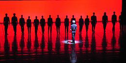 Tanzcompany, auf in rotes Licht getauchter Bühne, mit Choreograf im Vordergrund. 