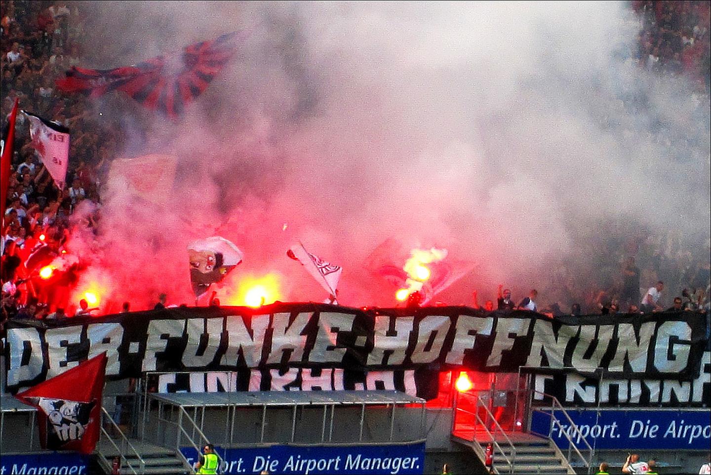 Fußballfans, zum Teil maskiert, werfen Böller und Rauchbomben. Auf der Tribüne brennt das Feuer.