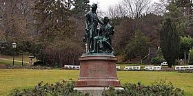 Zwei oxidierte Bronzestatuen von Joseph Lanner und Johann Strauß auf einem runden Fundament umringt von Ästen von Nadelbäumen, während im Hintergrund eine weite Wiese und Bäume zu sehen sind.