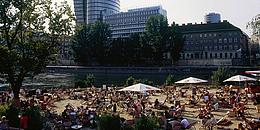 Viele Menschen sitzen auf Liegestühlen unter >Sonnenschirmen am Strand vorm Donaukanal