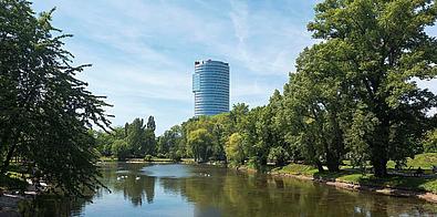 Ein spiegelndes Gewässer ist umringt von prächtigen grünen Bäumen an einem sonnigen Tag, während im Hintergrund der Florido Tower zu sehen ist.