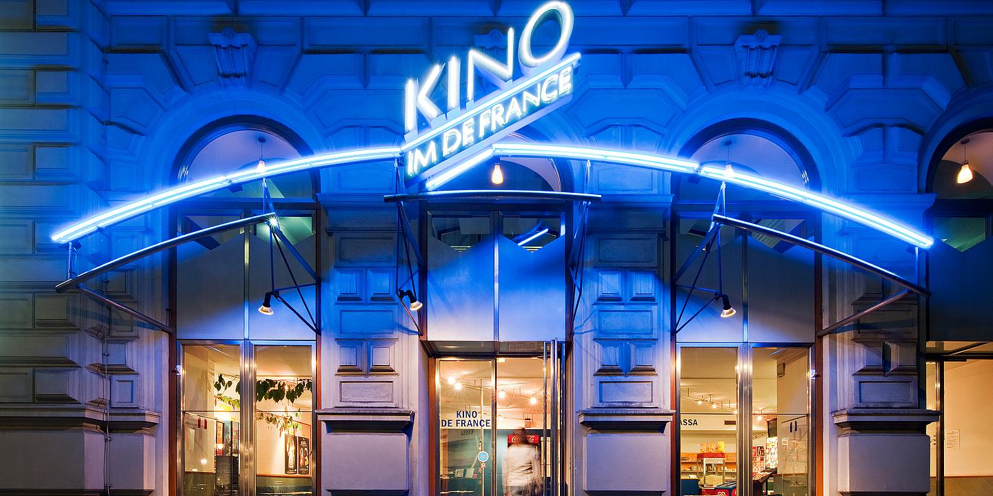 Frontalansicht auf das Kino De France bei Nacht von außen. Das Reklameschild des Kinos leutet Blau und man sieht in den hell beleuchteten Eingangsbereich hinein.