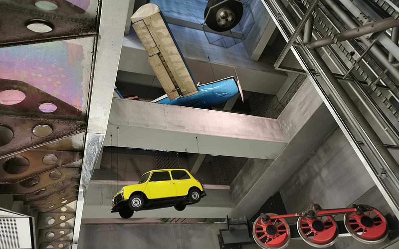 Ein gelber Mini Cooper, rote Räder einer Lokomotive, ein blaues Sportflugzeug und eine schwarze Mercury-Raumkapsel hängen zwischen Querstreben von der Decke