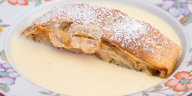 Apfelstrudel mit Schlagobers auf weißem Teller vor schwarzem Hintergrund