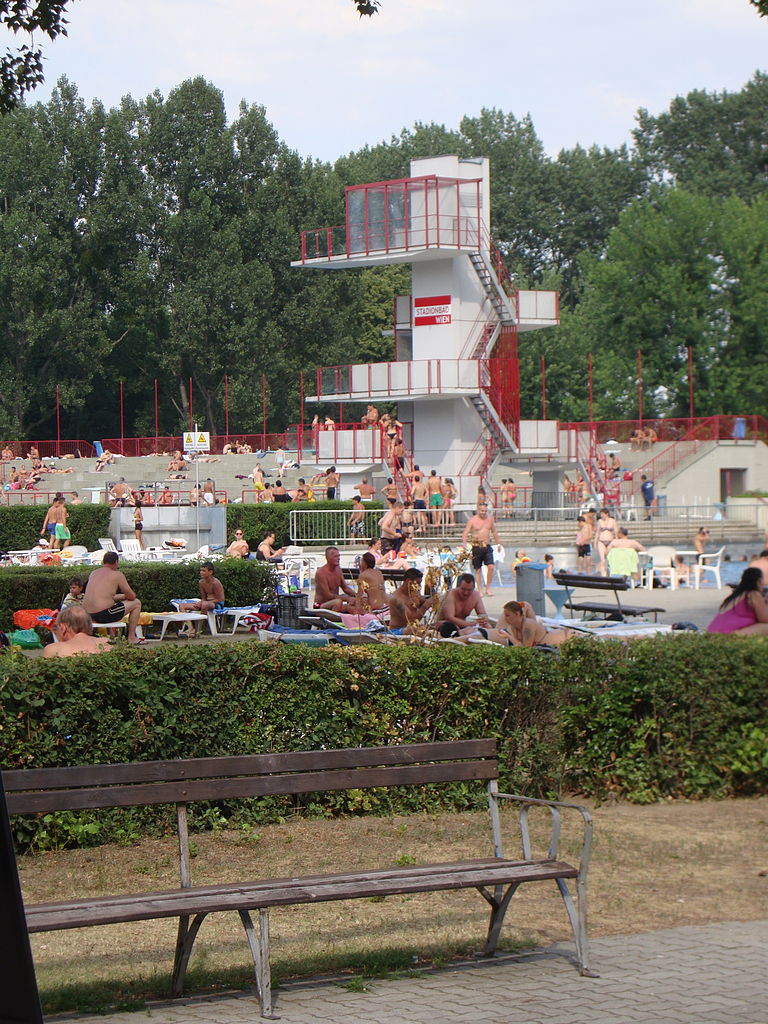 Ein 10m hoher Sprungturm in rot und weiß. Im vordergrund eine Parkbank.