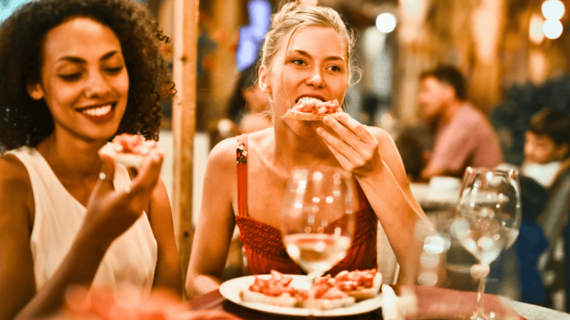 Zwei Frauen sitzen in einem Gastgarten und genießen Pizza und Wein