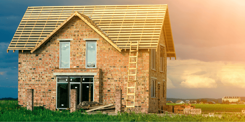 Ein Einfamilienhaus wird auf der grünen Wiese errichtet. Im Hintergrund geht die Sonne unter.