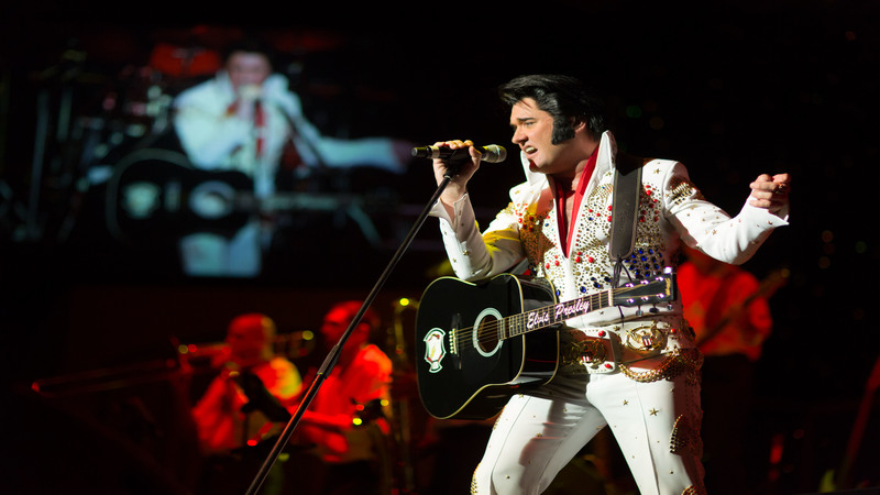 Elvis-Imitator in weißem Outfit singend auf der Bühne vor einer Videowall und einem Orchester im Hintergrund.